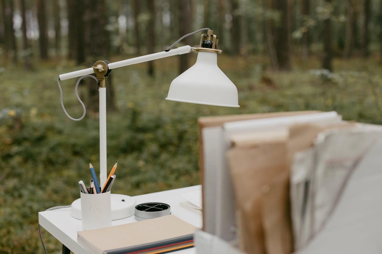 White desk lamp and books on a table in a lush forest. Ideal for nature-inspired workspaces.